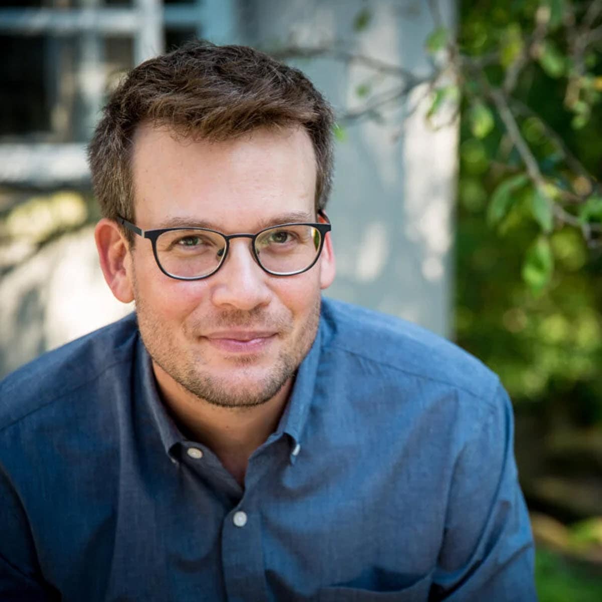 John Green smiling outdoors, wearing a blue shirt, glasses, and a beard