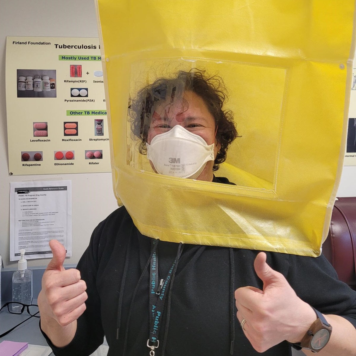 Moni Muraki wearing PPE in a clinical setting with their thumbs up wearing a black shirt