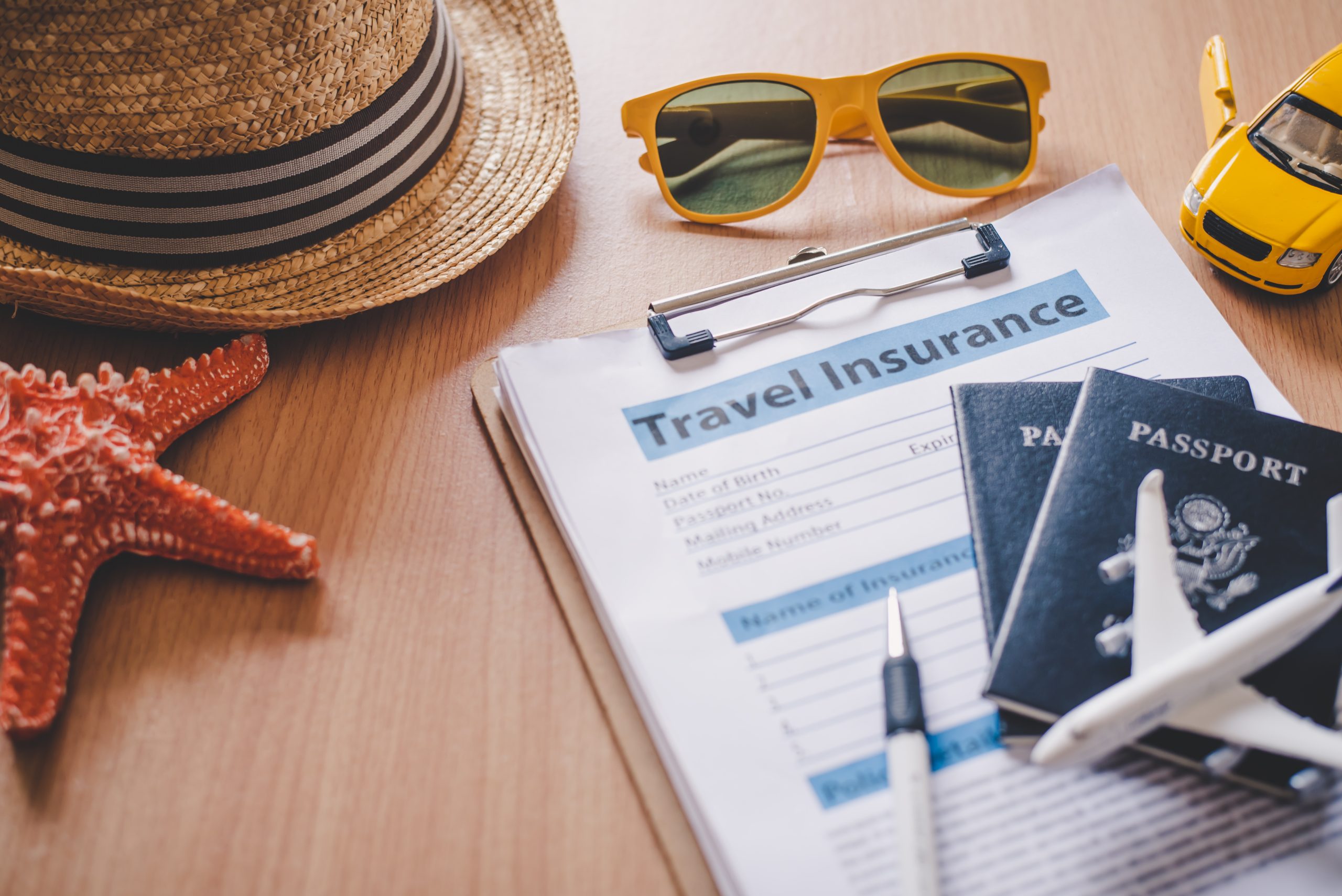 Clipboard with travel insurance documents, passports, hat, sunglasses, starfish, miniature airplane, and miniature car.