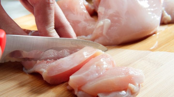 Unas manos cortando pollo crudo con un cuchillo.